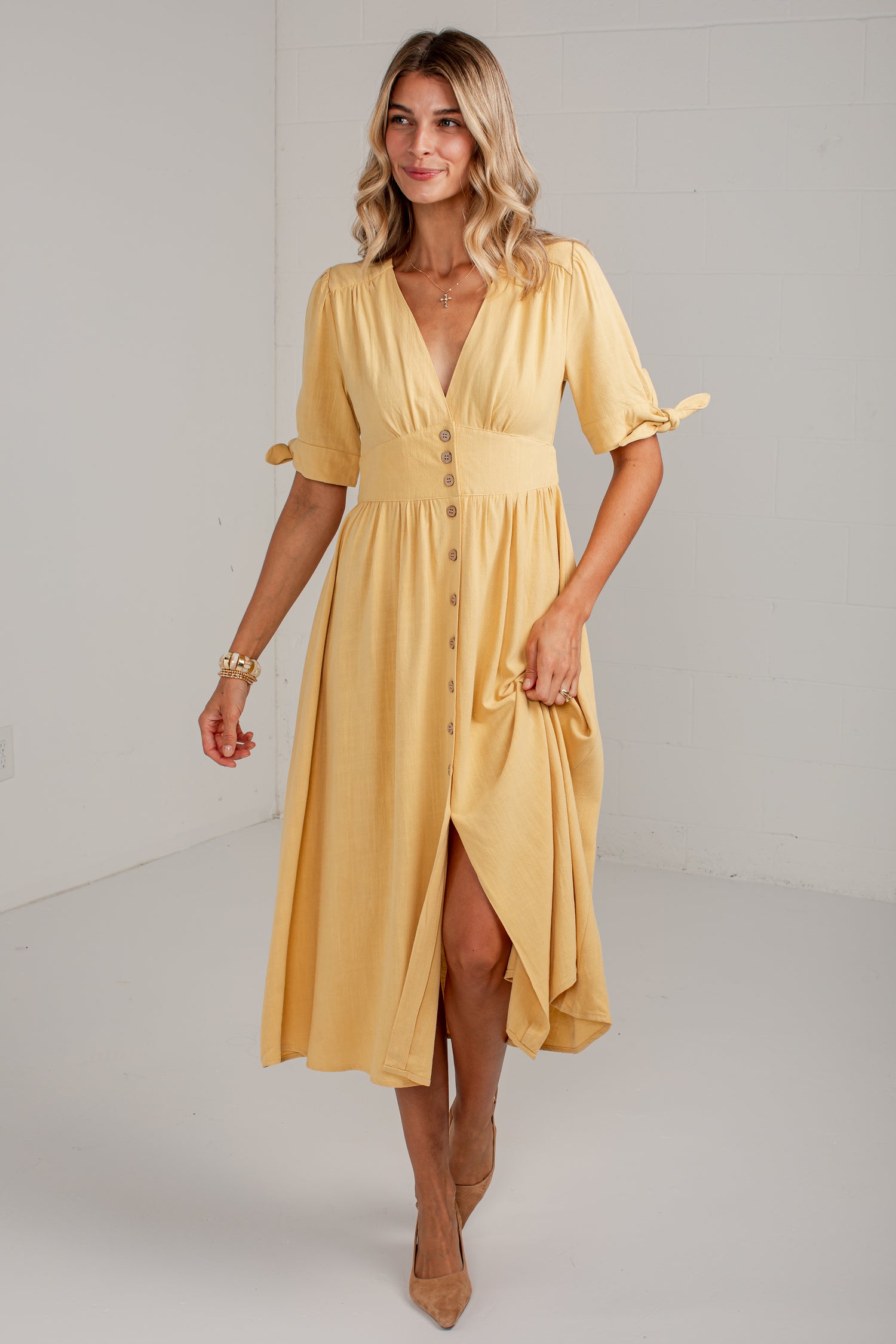 A woman modeling a mustard yellow midi dress with short flutter sleeves, a V-neckline, button-front detail, and a high-low hem, photographed in a bright white studio setting.