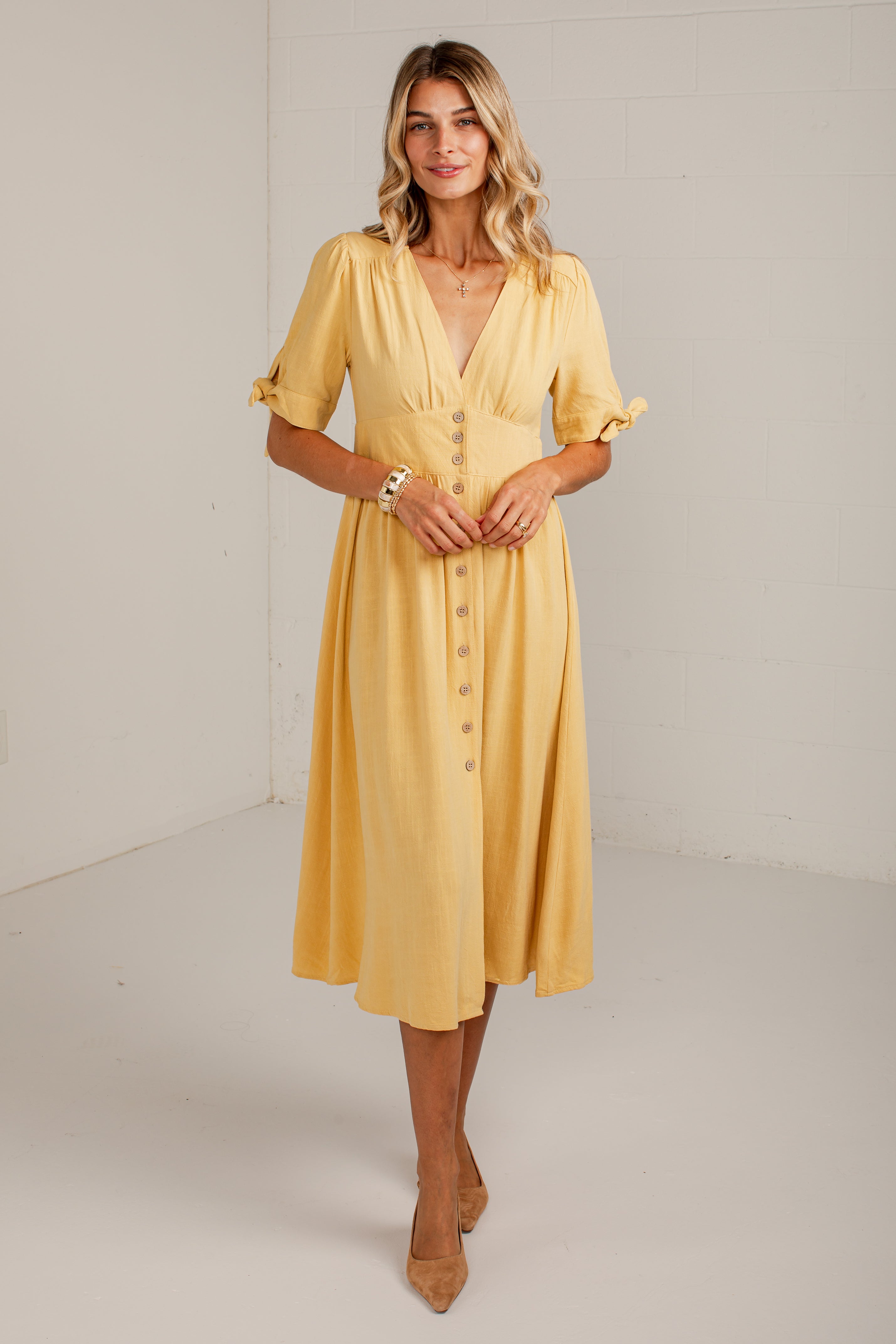 A woman modeling a yellow midi dress with short flutter sleeves, a V-neckline, and a button-front detail, standing in a bright studio setting.