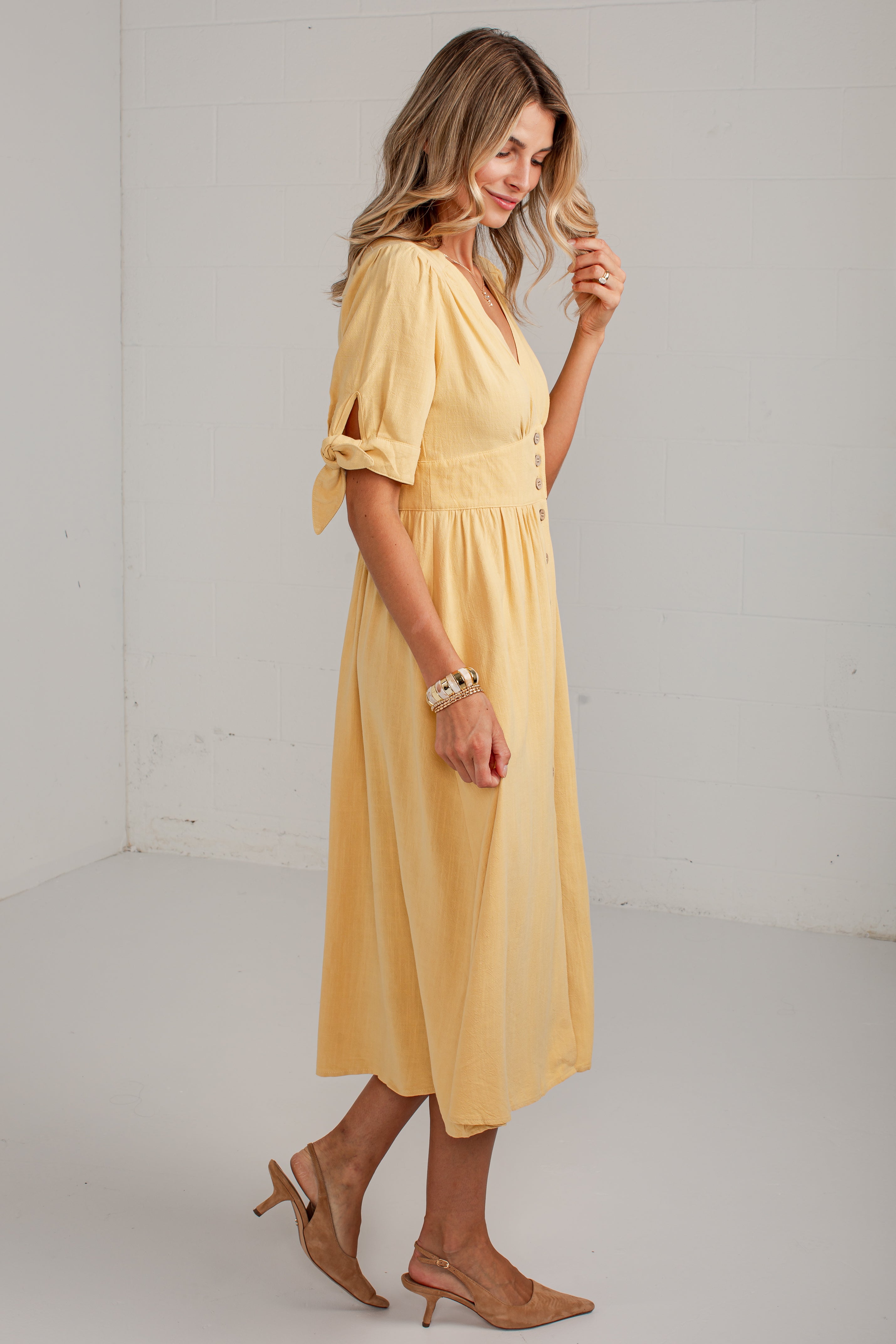 A woman modeling a light yellow midi dress with short sleeves, tie details at the cuffs, and a gathered waist, paired with tan heeled sandals in a bright studio setting.