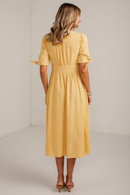 A woman wearing a yellow midi dress, shown from the back in a studio setting with a white brick wall background.