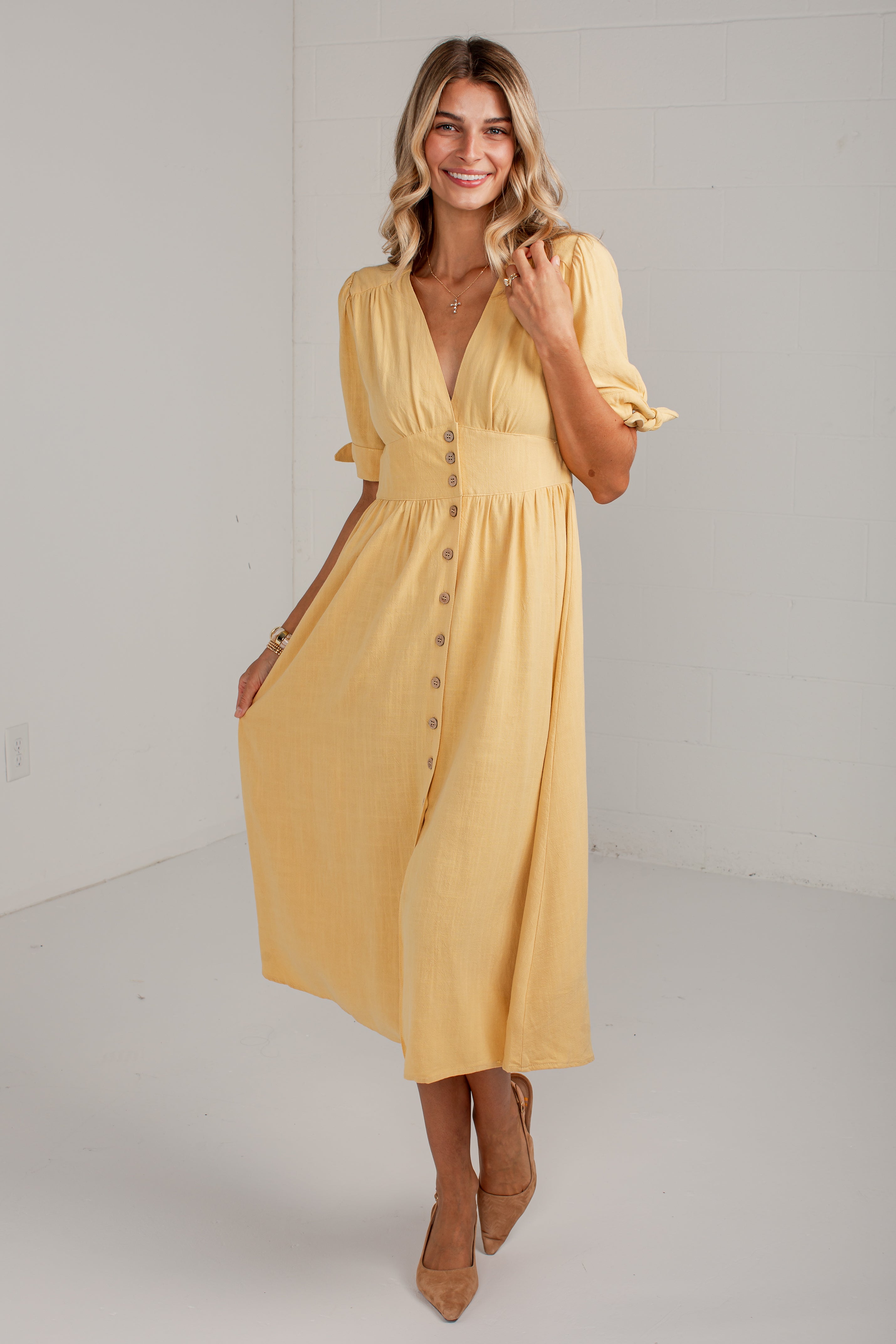 A woman modeling a yellow midi dress with puff sleeves, a V-neckline, and a button-down front, standing in a bright white studio setting.