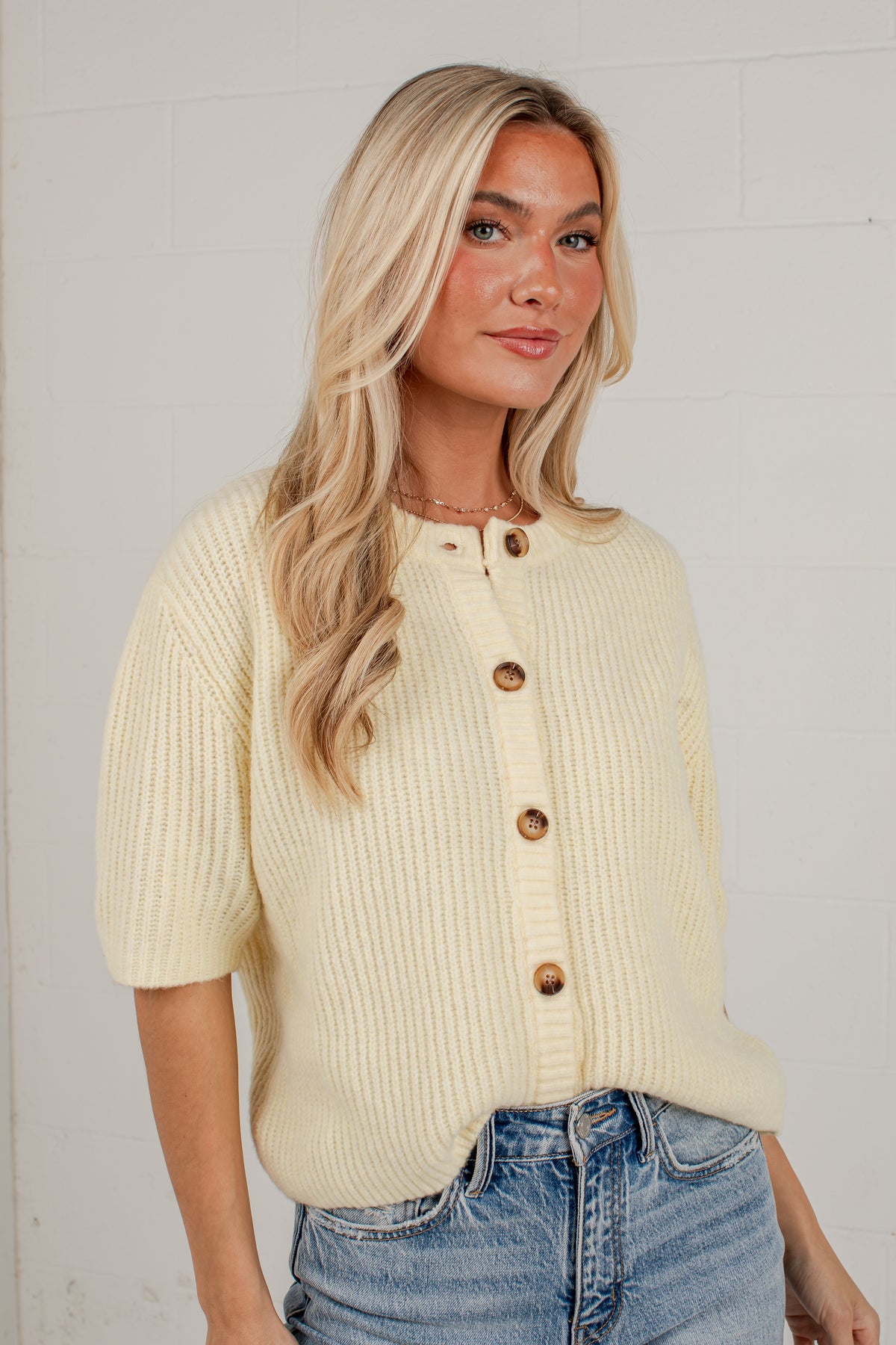 A blonde woman wearing a cream-colored ribbed knit cardigan with brown buttons and blue jeans, standing against a white brick wall.