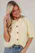A woman wearing a cream-colored ribbed knit short-sleeve cardigan with brown buttons, paired with blue jeans, posing against a white brick wall.