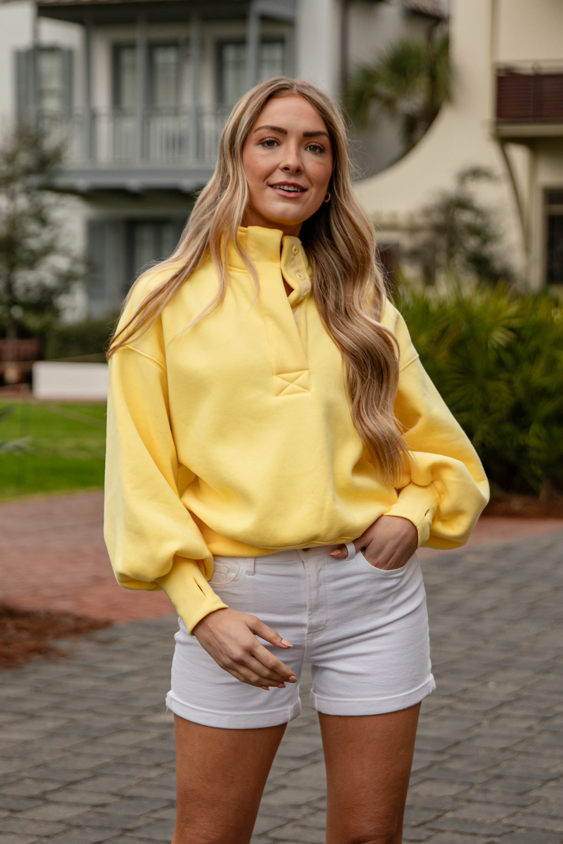 Woman wearing a yellow sweatshirt and white shorts standing outdoors.