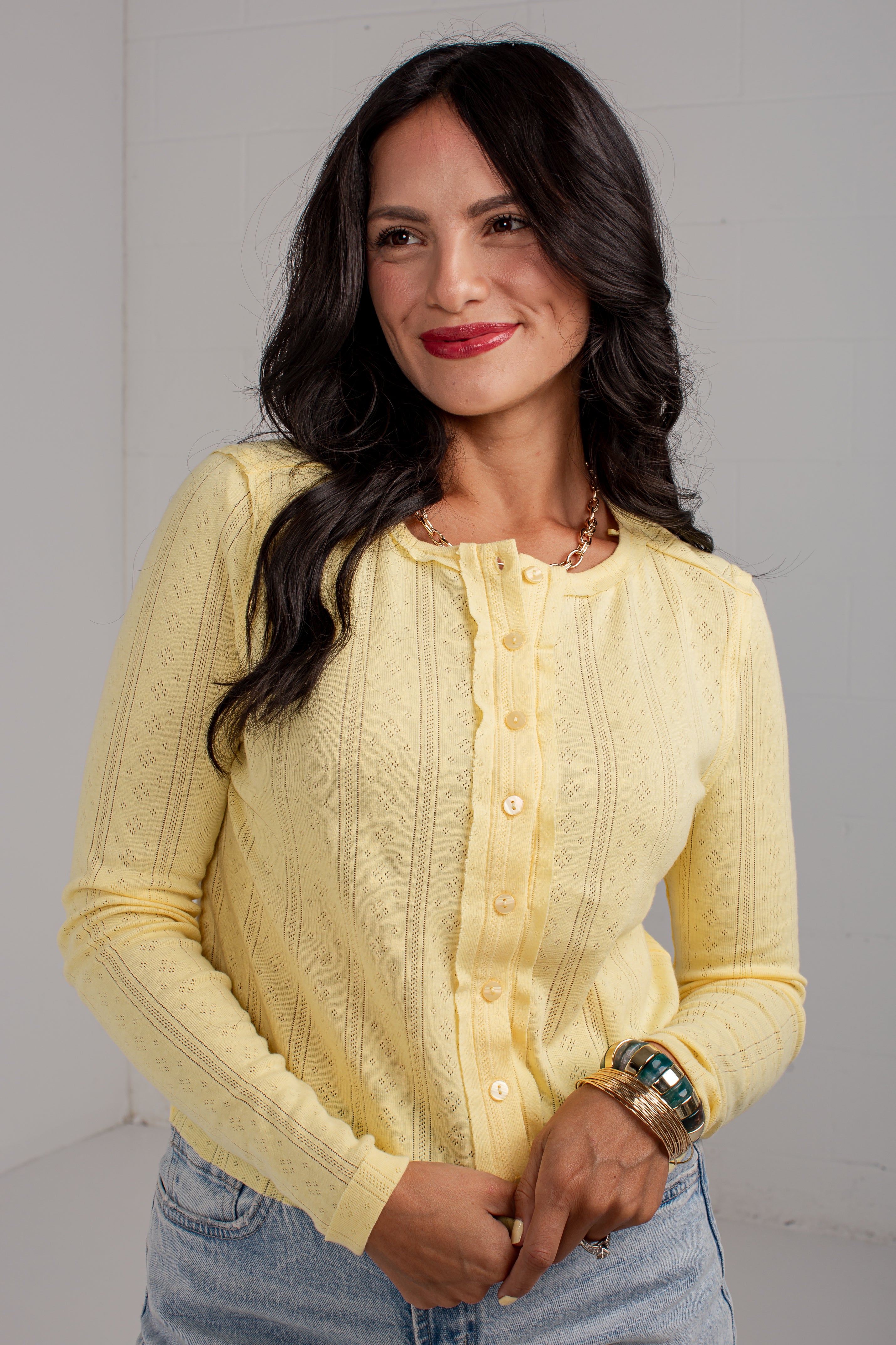 A woman wearing a pale yellow textured knit button-up cardigan sweater and light blue jeans, standing against a light gray wall.