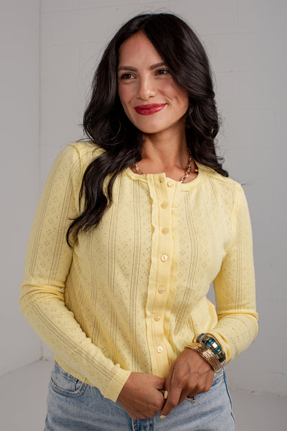 A woman wearing a pale yellow textured knit button-up cardigan sweater and light blue jeans, standing against a light gray wall.