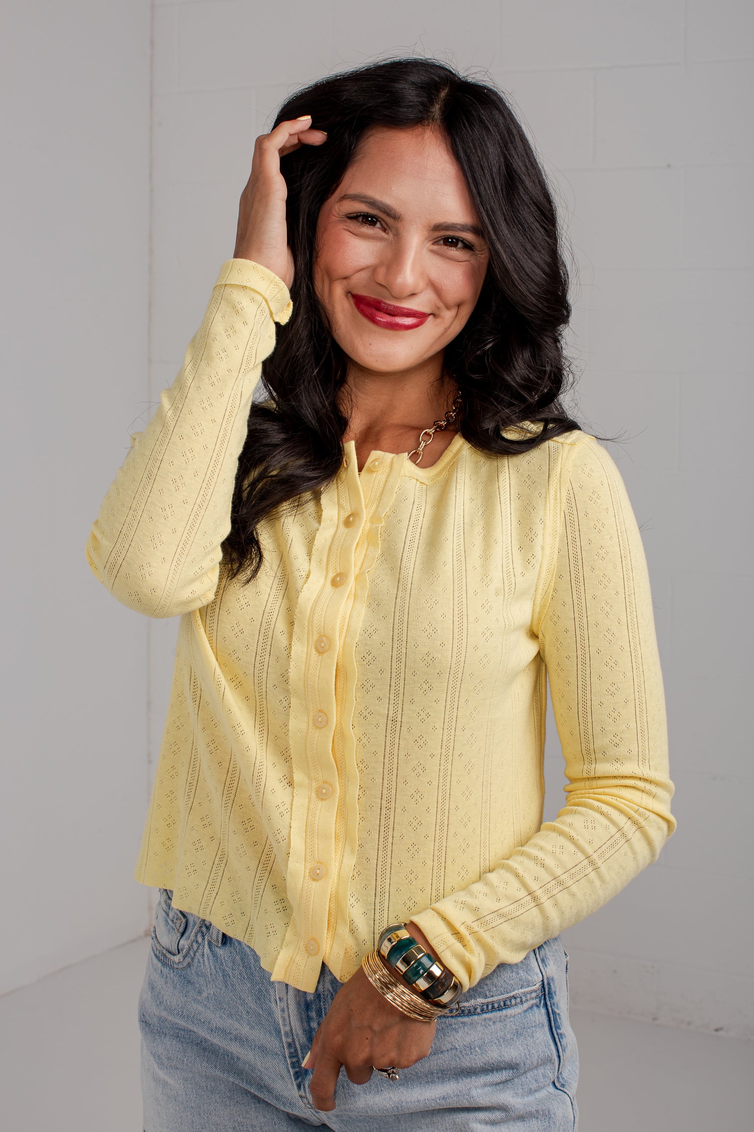 A woman wearing a pale yellow long-sleeve button-front cardigan with textured knit details, paired with light-wash blue jeans, standing against a light gray wall.