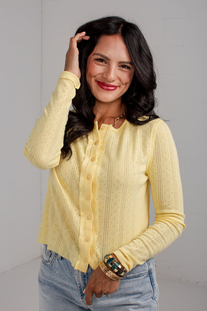 A woman wearing a pale yellow long-sleeve button-front cardigan with textured knit details, paired with light-wash blue jeans, standing against a light gray wall.