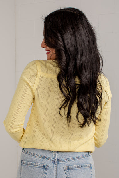 A woman with long, wavy dark hair is shown from behind, wearing a pale yellow textured knit long-sleeve top and light blue jeans against a white paneled wall.