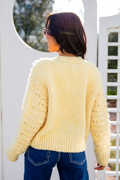 Woman wearing a yellow knitted sweater and blue jeans standing in front of a white lattice fence.