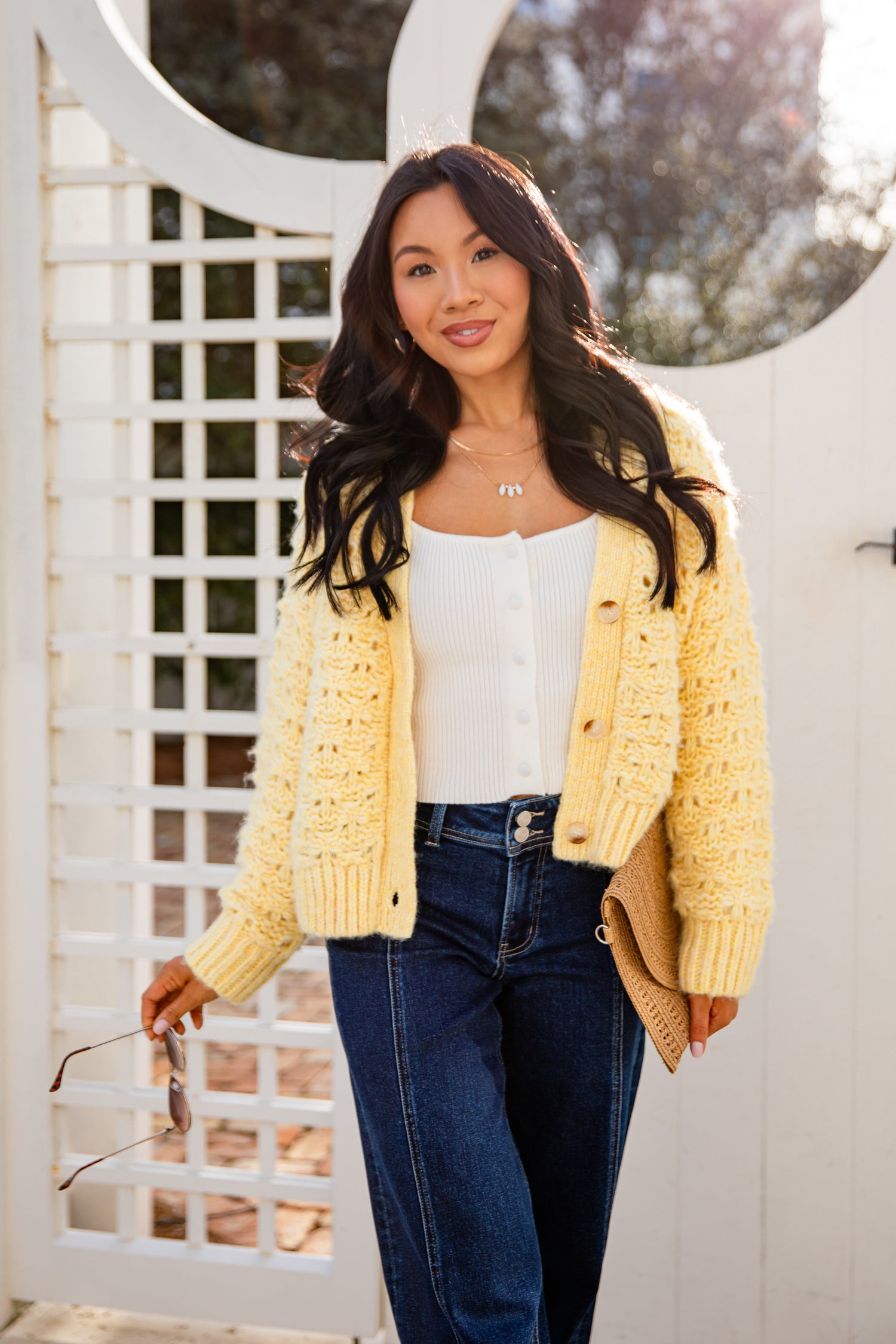 Woman wearing a yellow cardigan over a white top and blue jeans, standing in front of a white lattice fence.