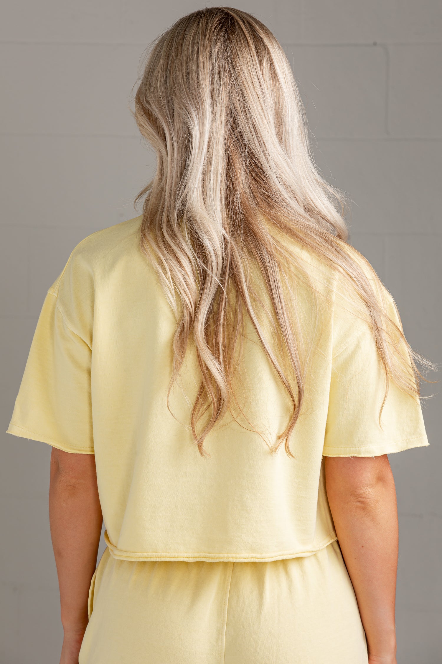 A woman viewed from behind wearing a pale yellow short-sleeve cropped top and matching bottoms against a light gray wall.
