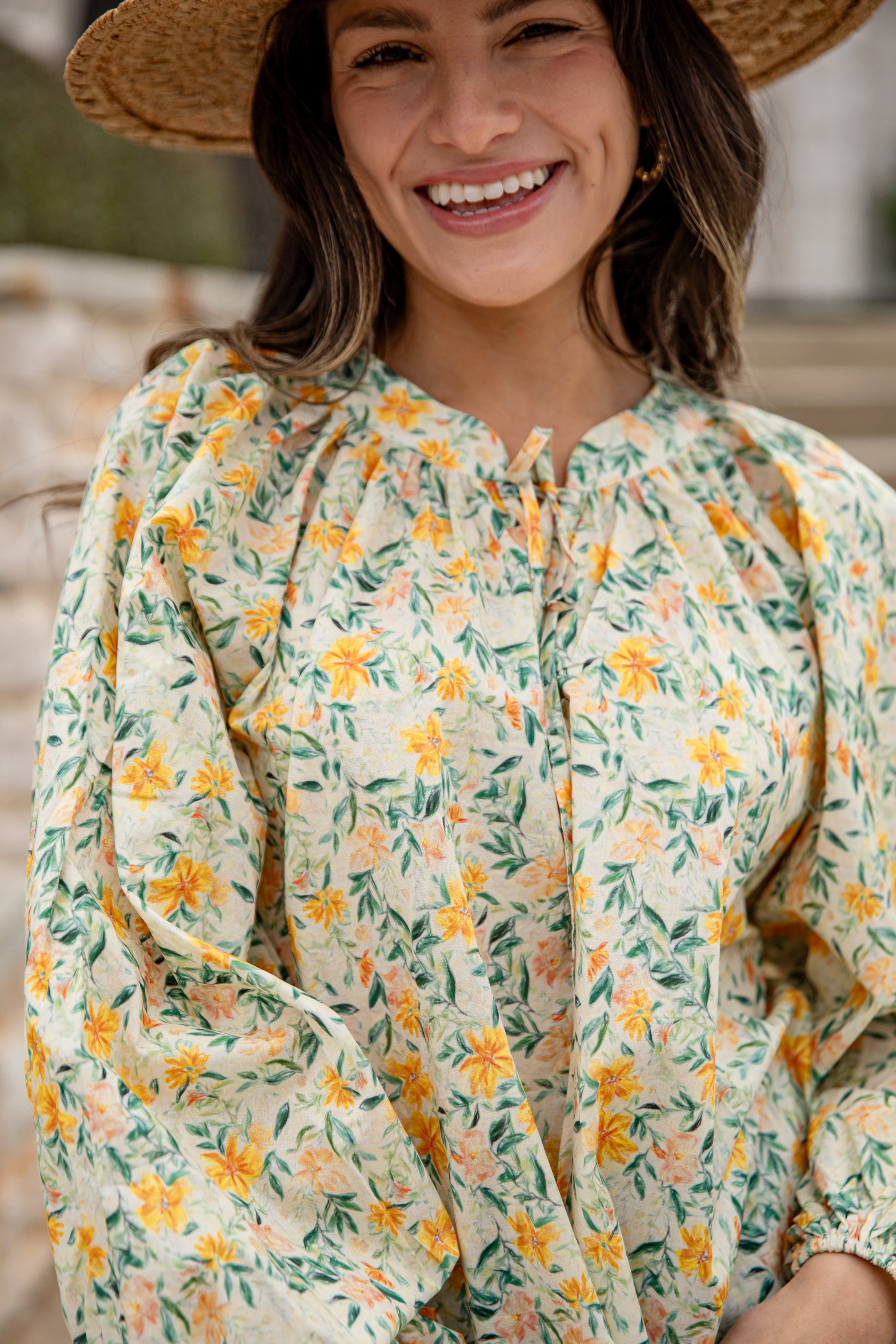 Woman wearing a floral blouse with a straw hat, smiling outdoors.