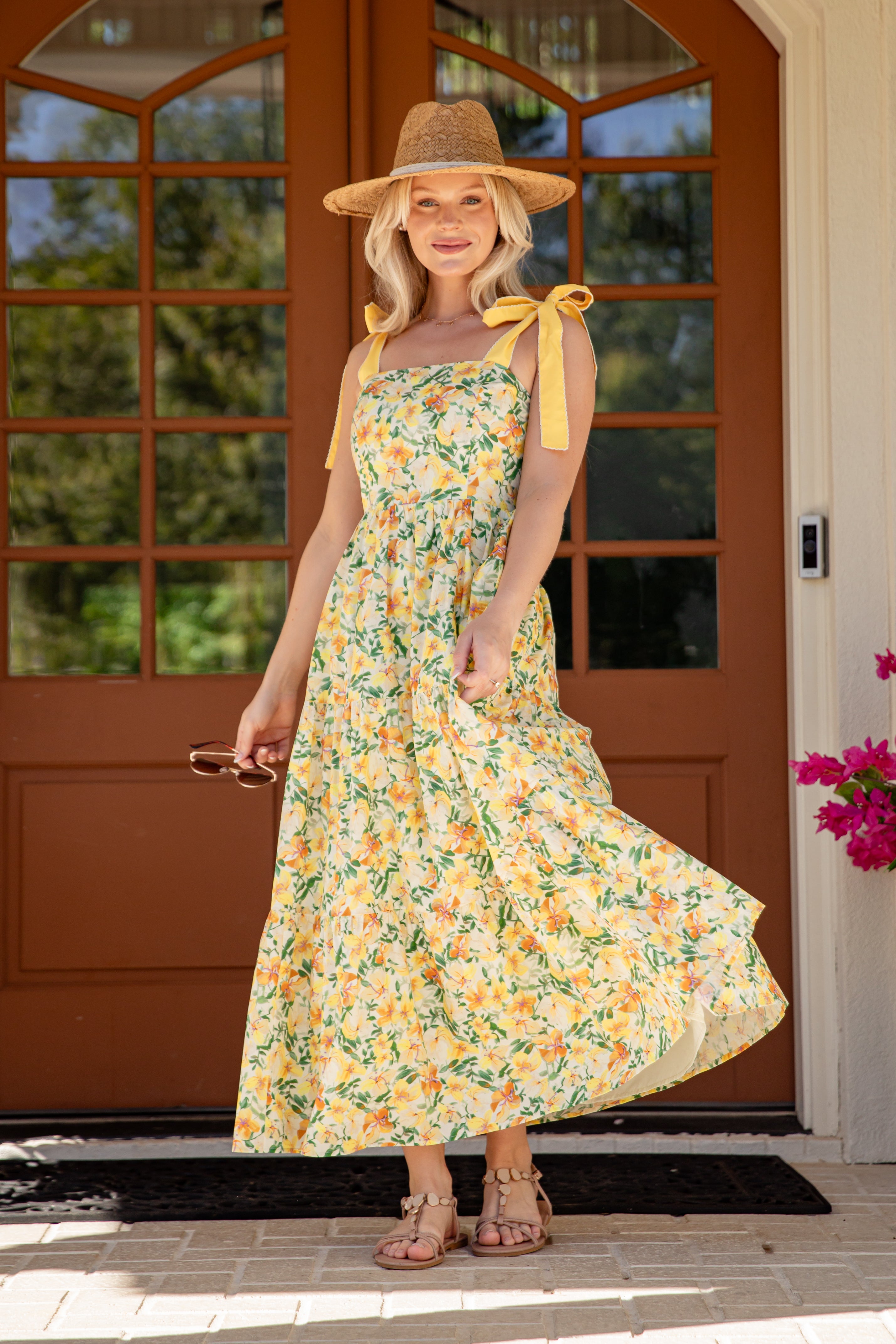 Woman in a floral dress and hat standing in front of a brown door with greenery.