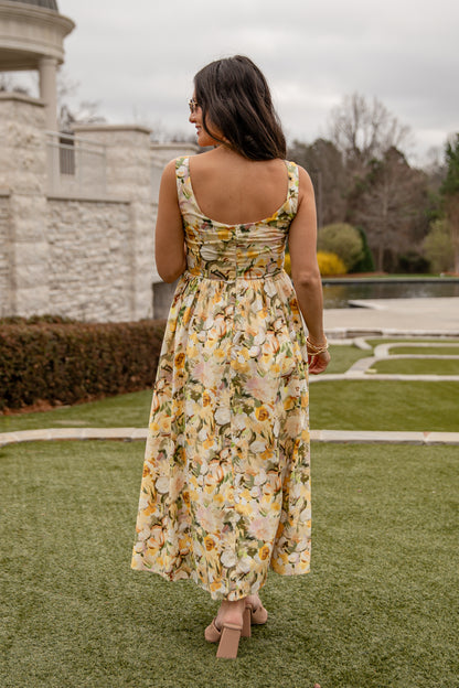 Woman in a floral dress standing on grass with a stone wall and trees in the background