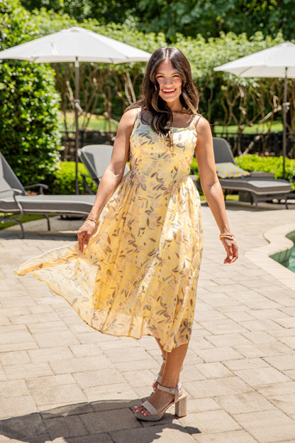 Woman in a yellow dress standing outdoors on a patio with greenery and umbrellas in the background.