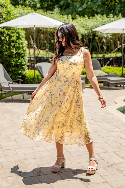 Woman in a yellow floral dress standing outdoors on a patio with greenery and lounge chairs.