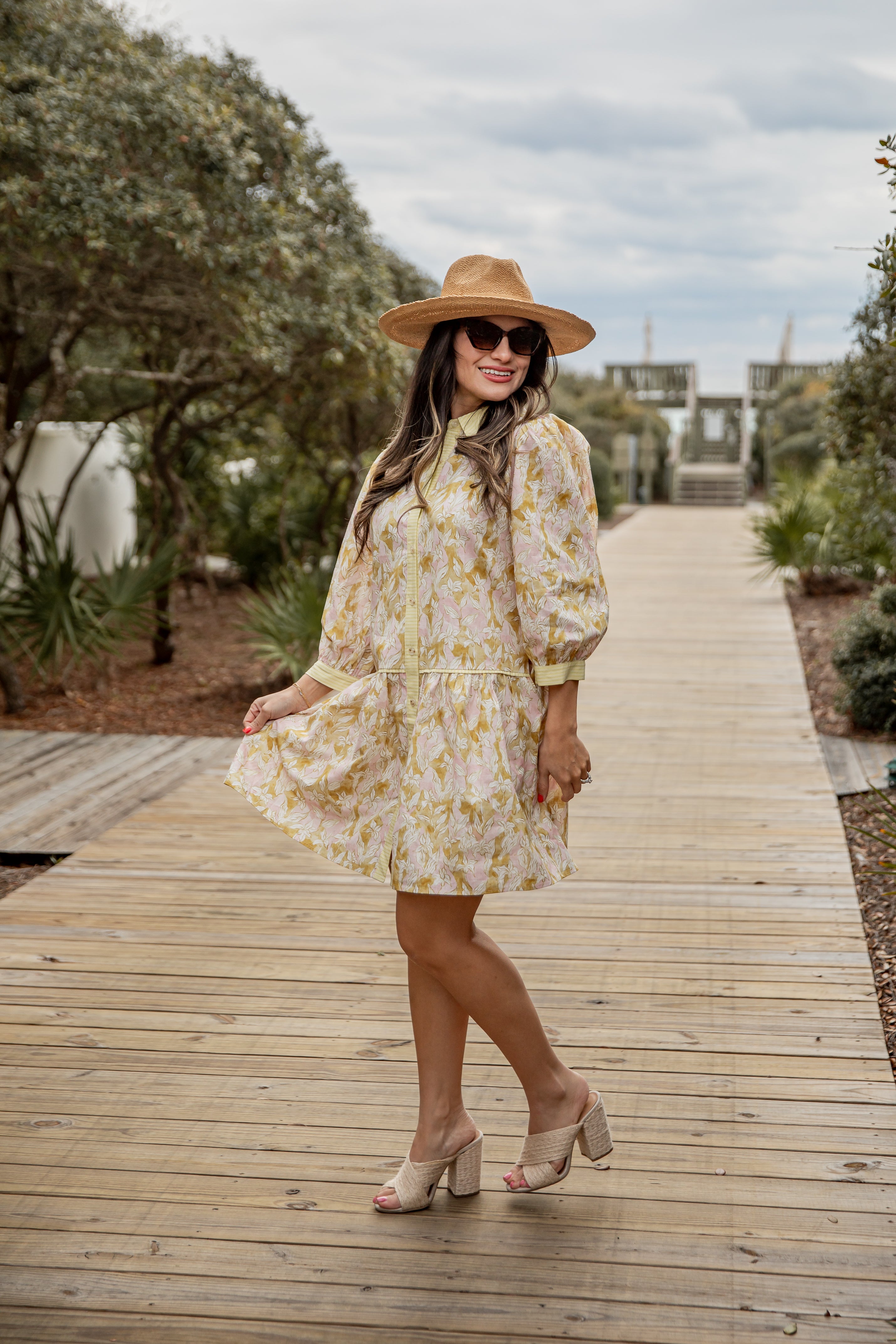 Woman in a floral dress and hat standing on a wooden pathway with trees and sky in the background