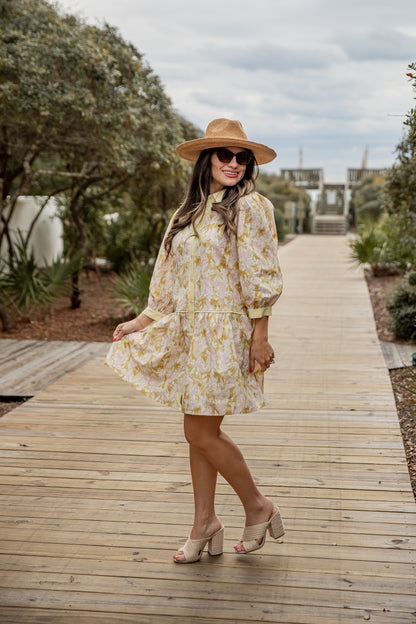 Woman in a floral dress and hat standing on a wooden pathway with trees and sky in the background
