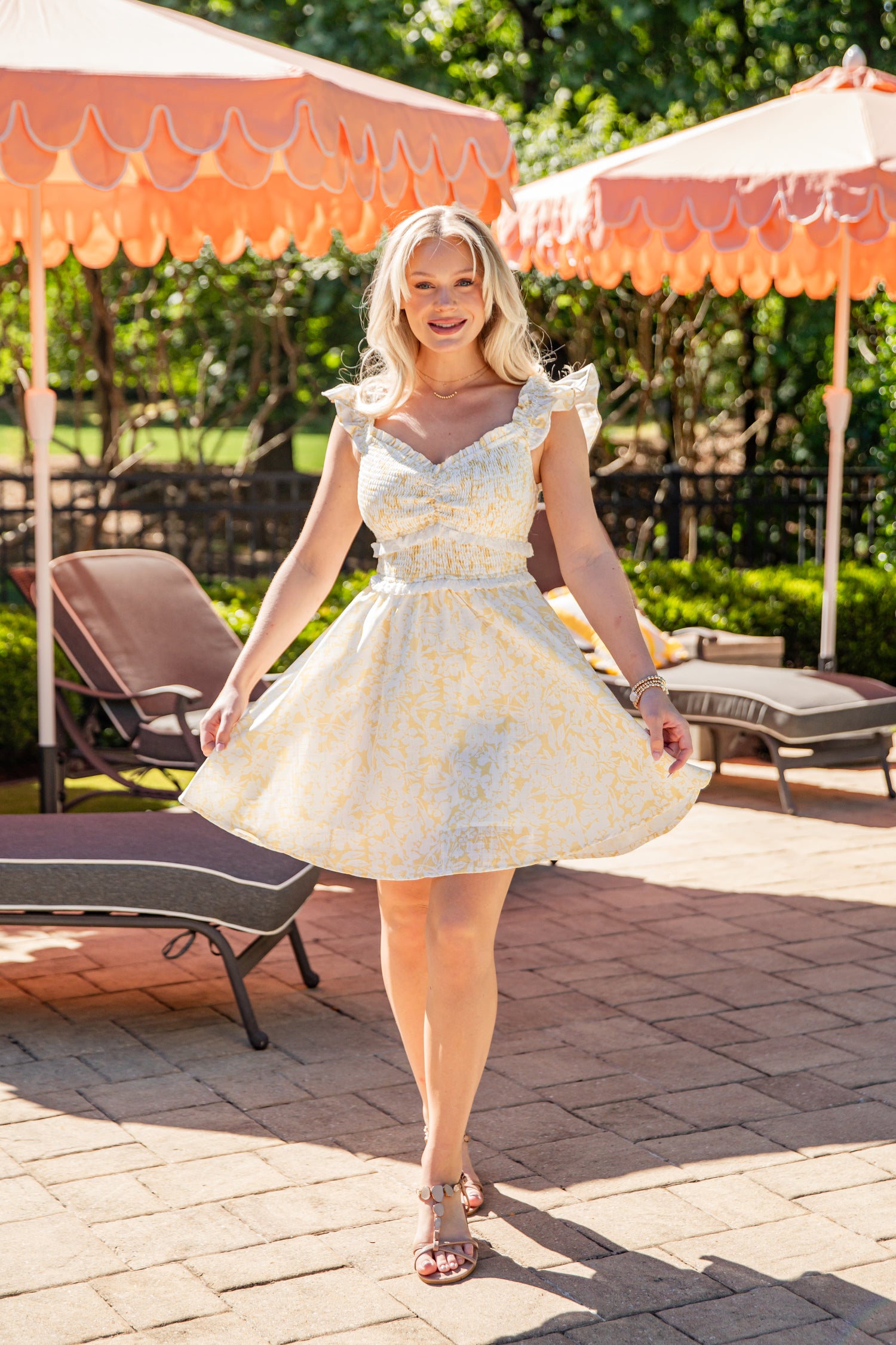 Woman in a white floral dress standing outdoors with orange umbrellas and lounge chairs.