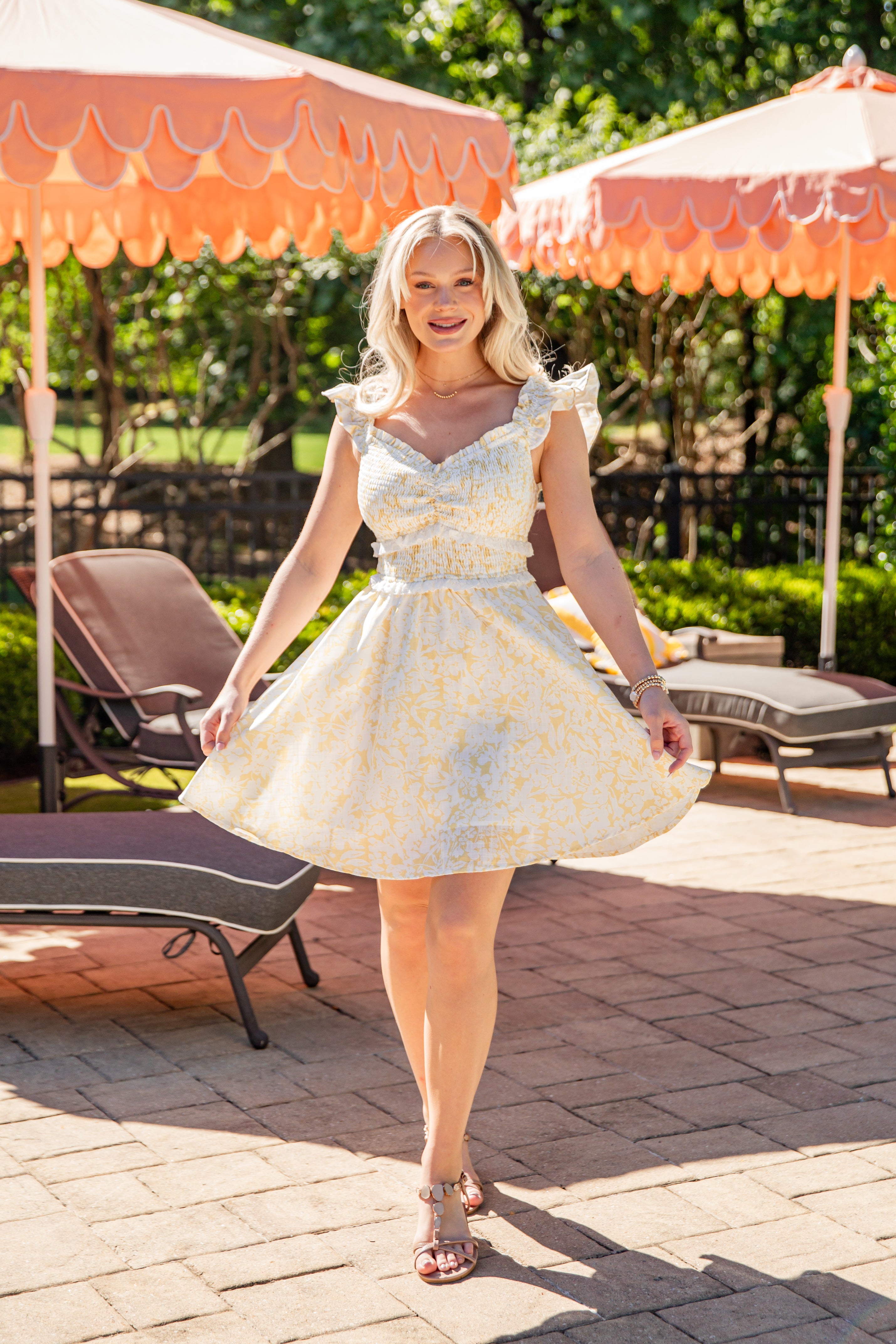 Woman in a white floral dress standing outdoors with orange umbrellas and lounge chairs.