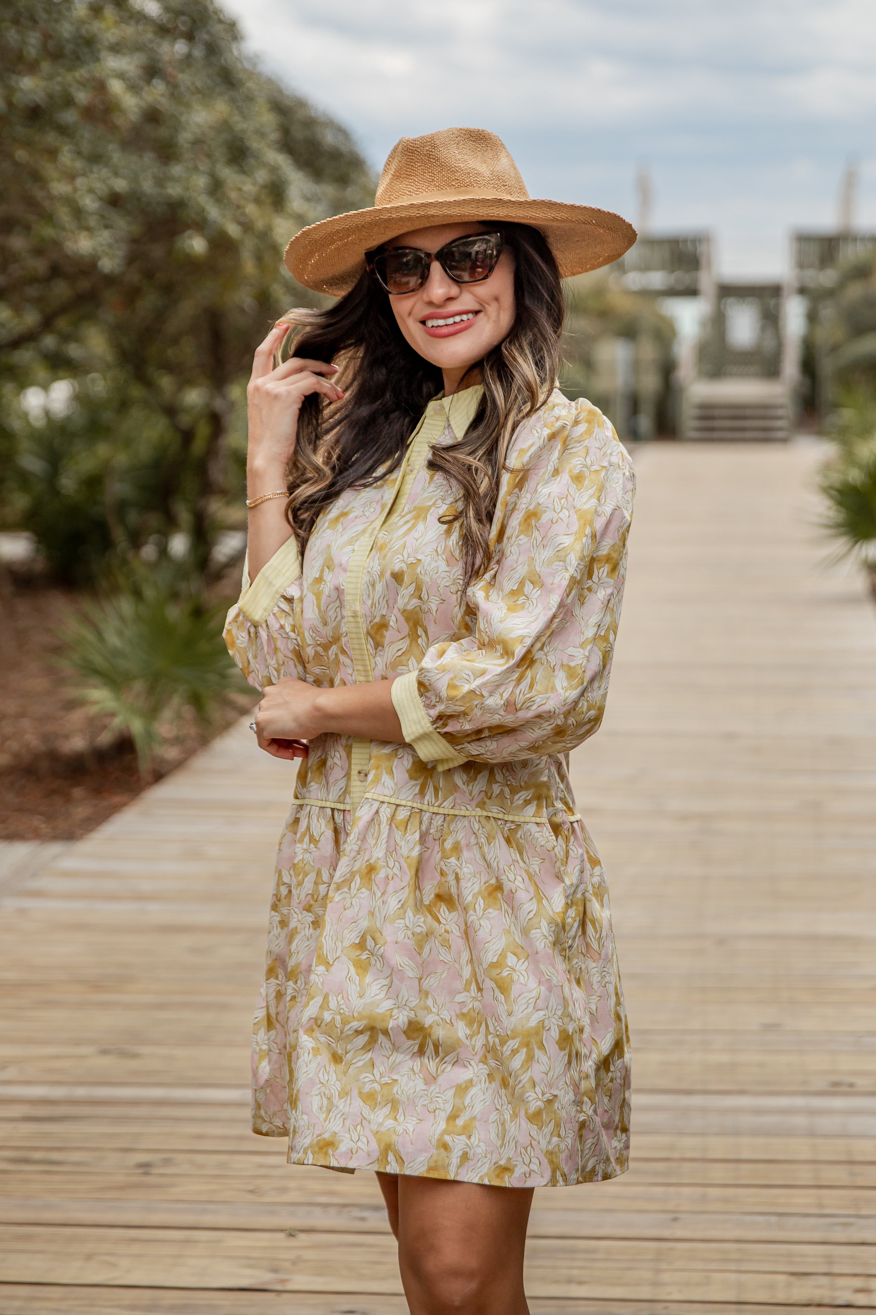Woman in a floral dress and hat standing on a wooden boardwalk with trees in the background