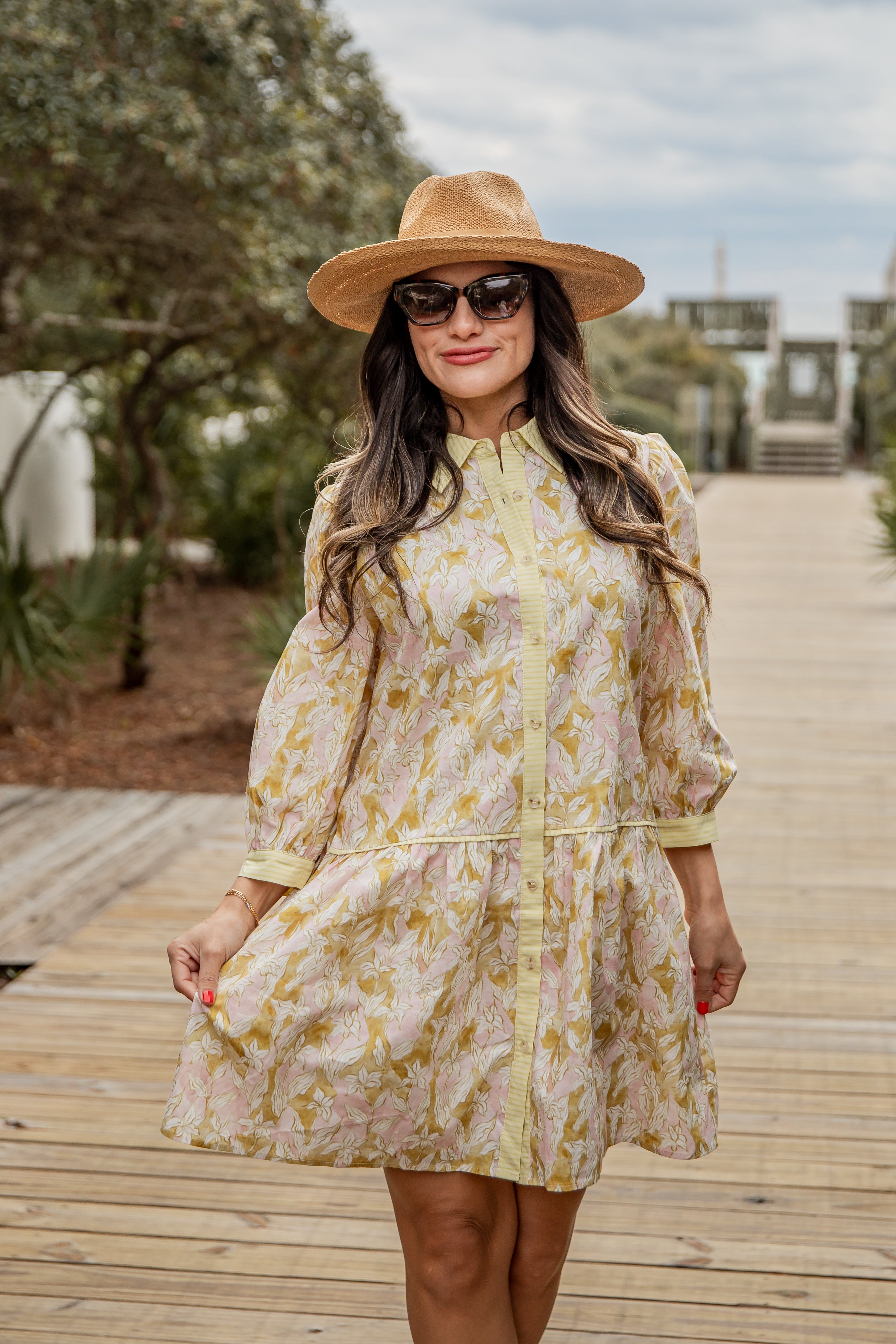 Woman in a floral dress and hat standing on a wooden boardwalk with trees and sky in the background
