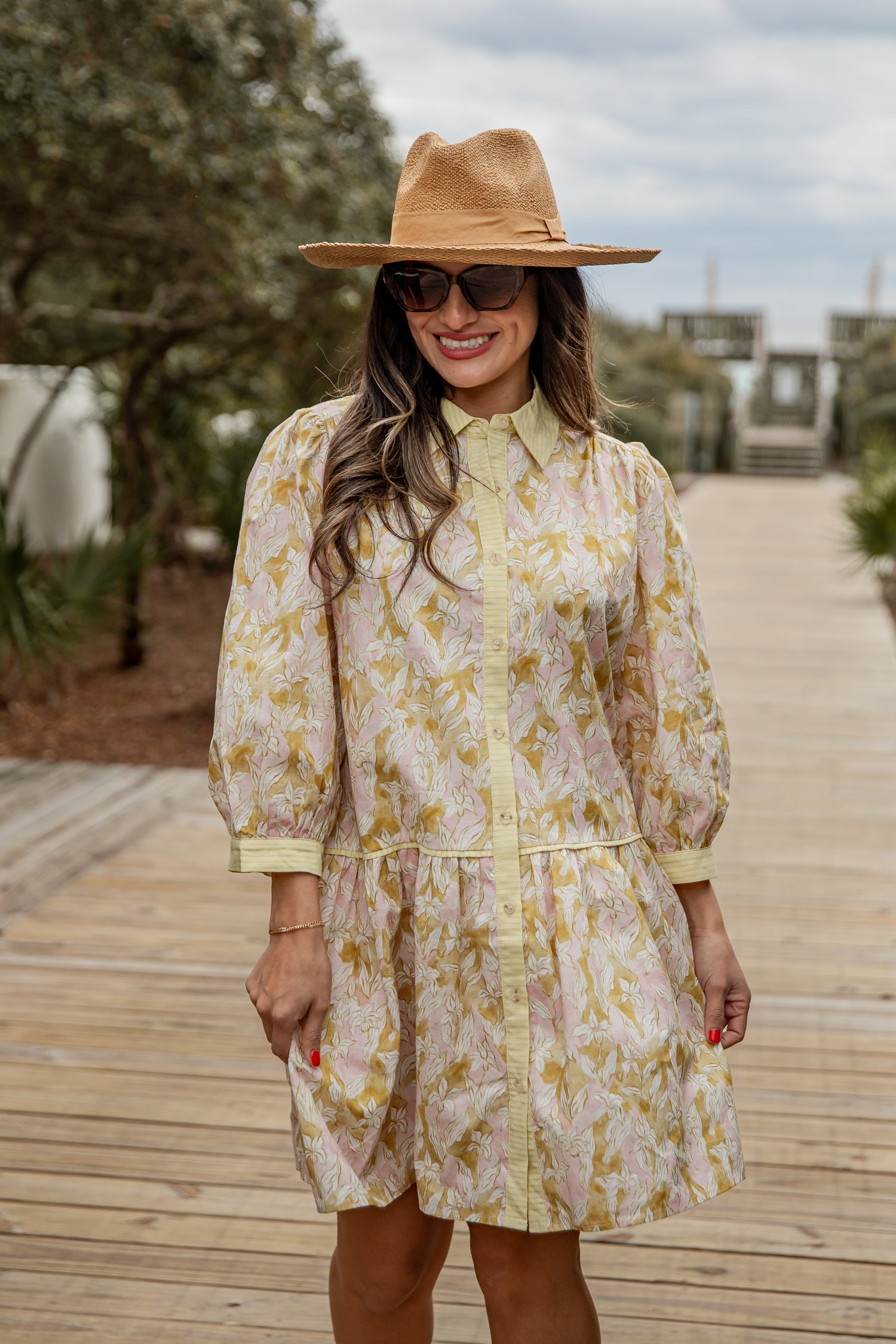 Woman wearing a floral dress and hat on a wooden pathway with greenery in the background