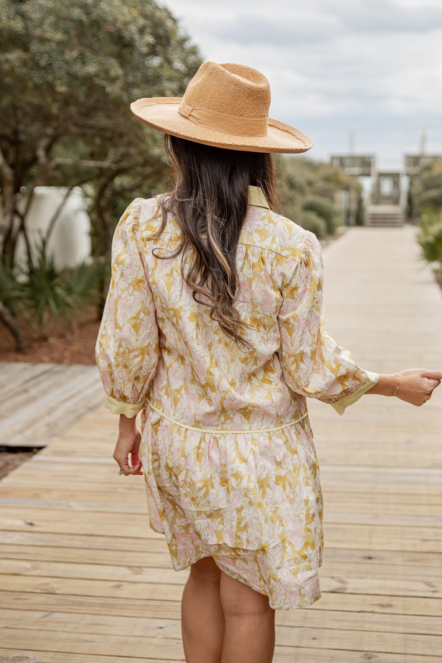 Woman in a floral dress and straw hat walking on a wooden path outdoors.