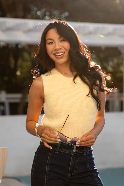 Woman wearing a yellow sleeveless top and black pants, holding sunglasses, with a blurred outdoor background.