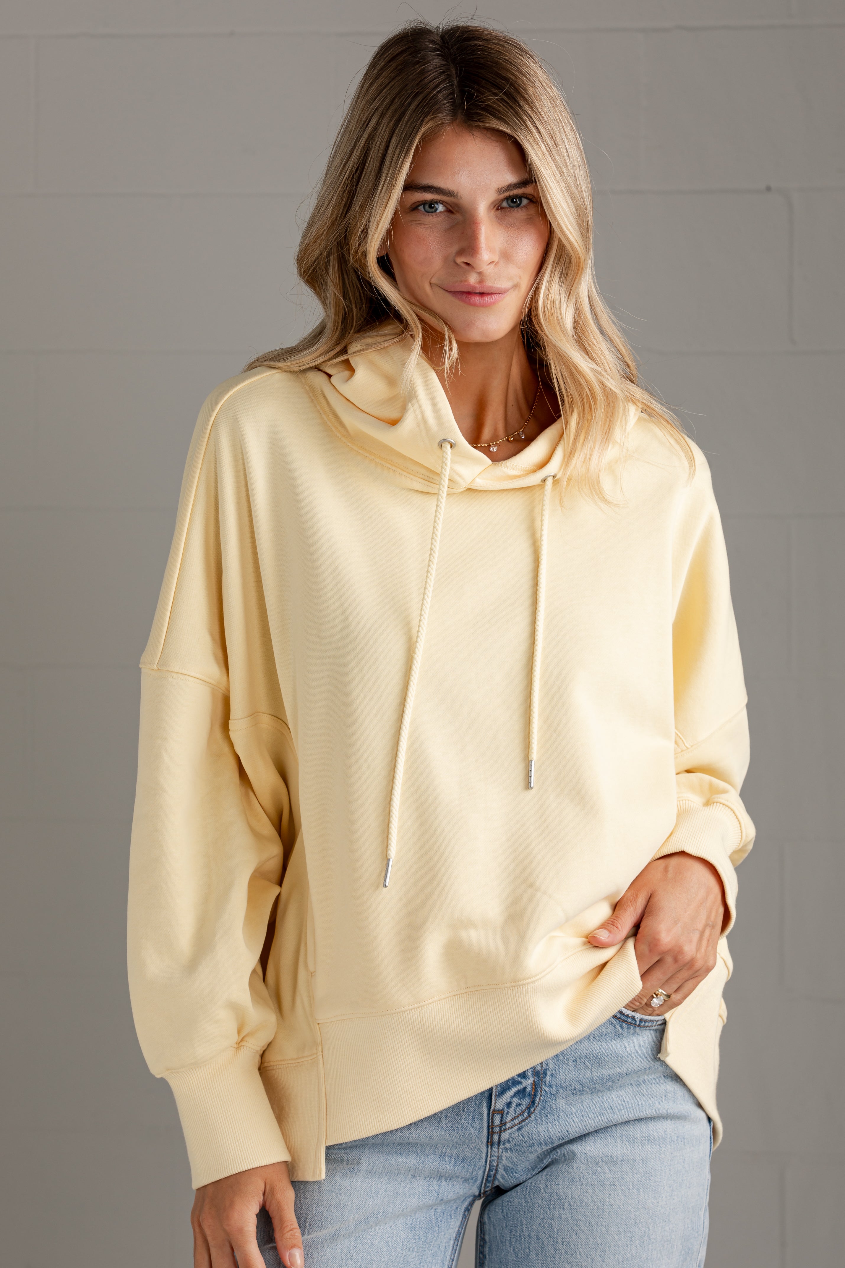 A woman wearing a pale yellow oversized pullover hoodie with drawstrings and front pouch pockets, paired with light blue jeans, standing against a gray paneled wall.