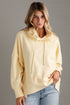 A woman wearing a pale yellow oversized pullover hoodie with drawstrings and front pouch pockets, paired with light blue jeans, standing against a gray paneled wall.