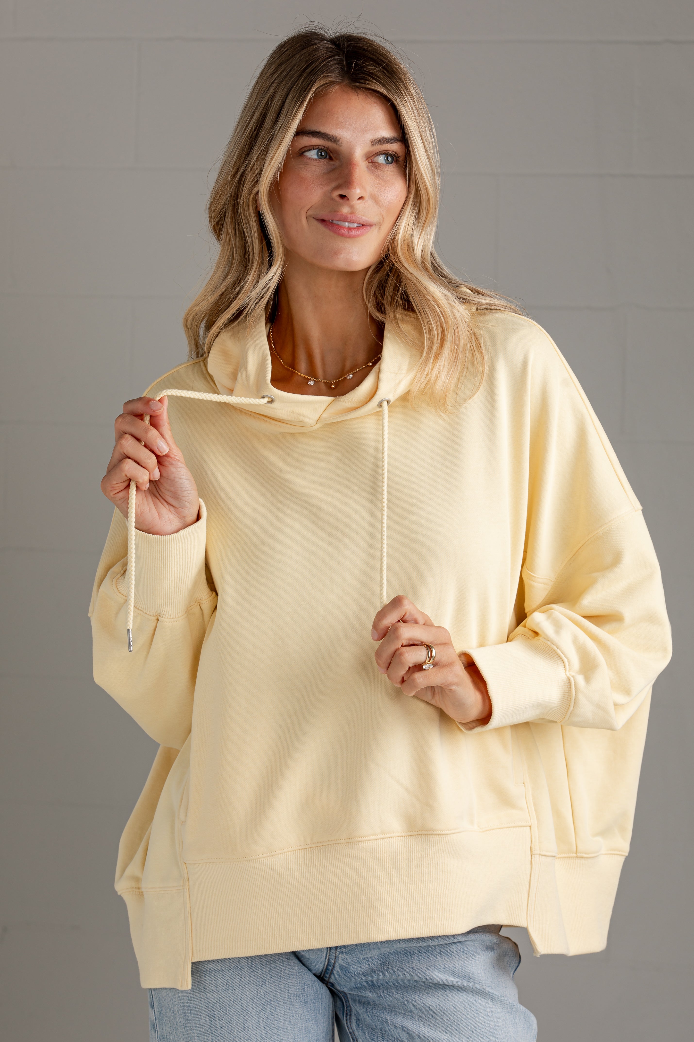 A woman wearing a pale yellow oversized pullover hoodie with drawstrings and front pouch pocket, paired with light blue jeans, standing against a gray tiled wall.