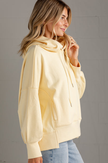 A woman wearing a pale yellow oversized pullover hoodie with drawstrings and front pockets, paired with light blue jeans, standing against a gray brick wall.