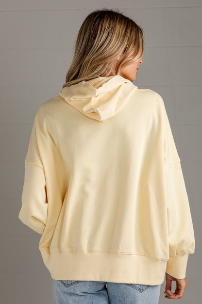 A woman viewed from the back wearing an oversized pale yellow hoodie and light blue jeans, standing against a light gray paneled wall.