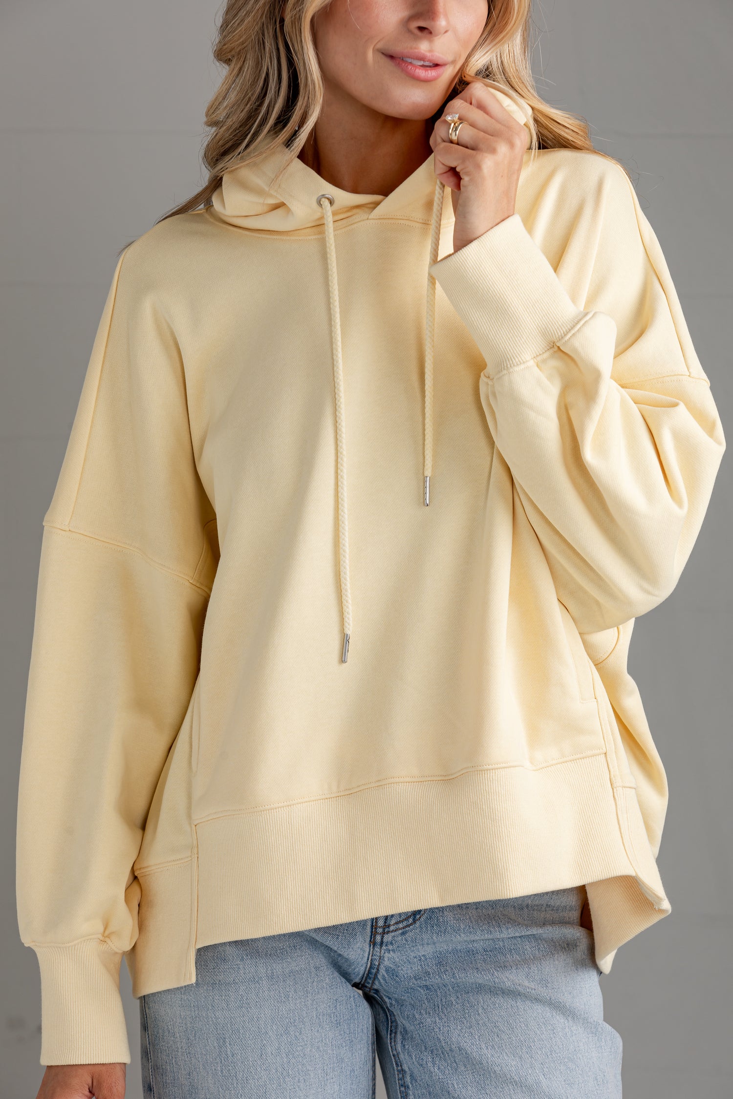 A woman wearing a pale yellow oversized pullover hoodie with drawstrings and a front kangaroo pocket, paired with light blue jeans, against a neutral gray background.