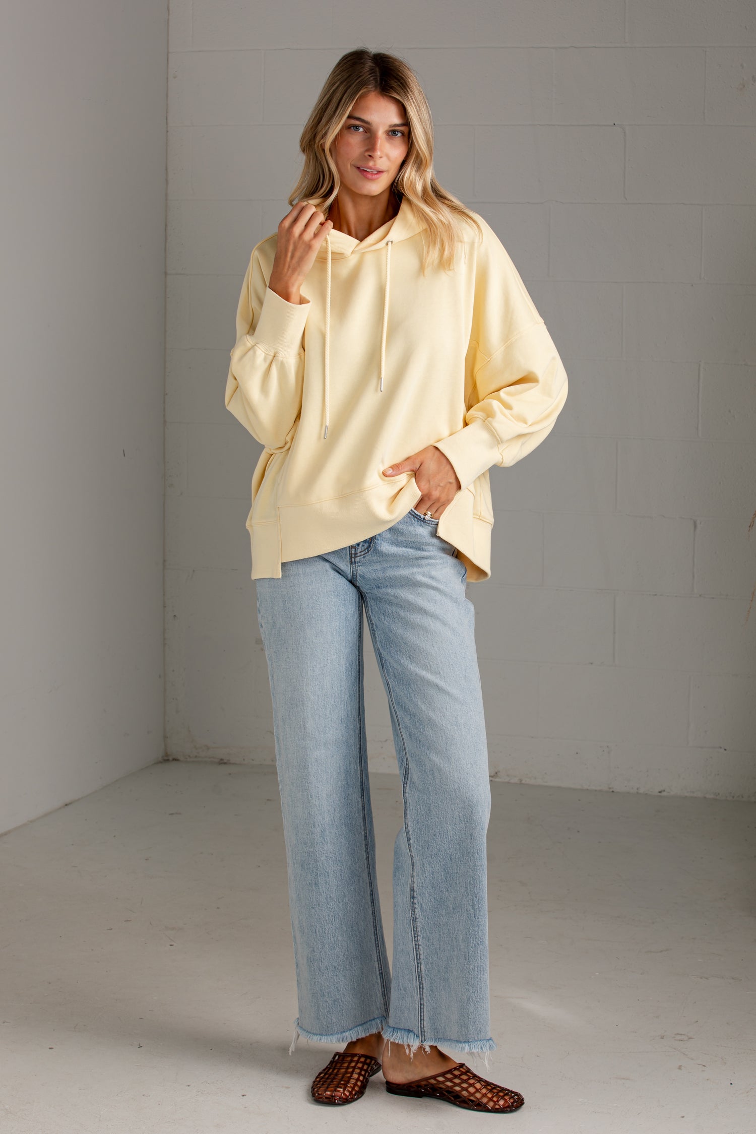 A woman modeling an oversized pale yellow hoodie with light-wash blue jeans and brown woven mule flats, standing in a minimalist gray studio corner.