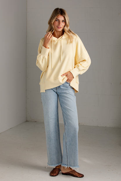 A woman modeling an oversized pale yellow hoodie with light-wash blue jeans and brown woven mule flats, standing in a minimalist gray studio corner.