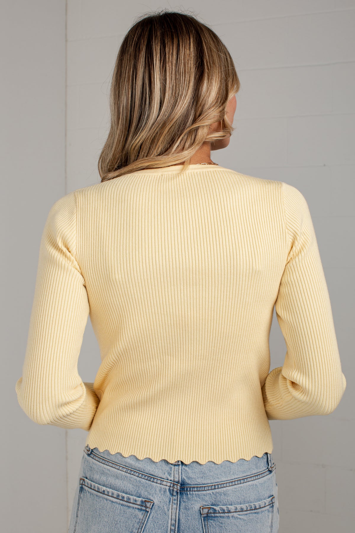 A woman viewed from behind wearing a pale yellow ribbed knit long-sleeve top with a scalloped hem and light blue jeans against a light gray wall.