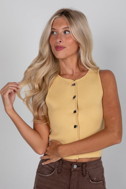 Woman wearing a yellow sleeveless top with black buttons on a plain background