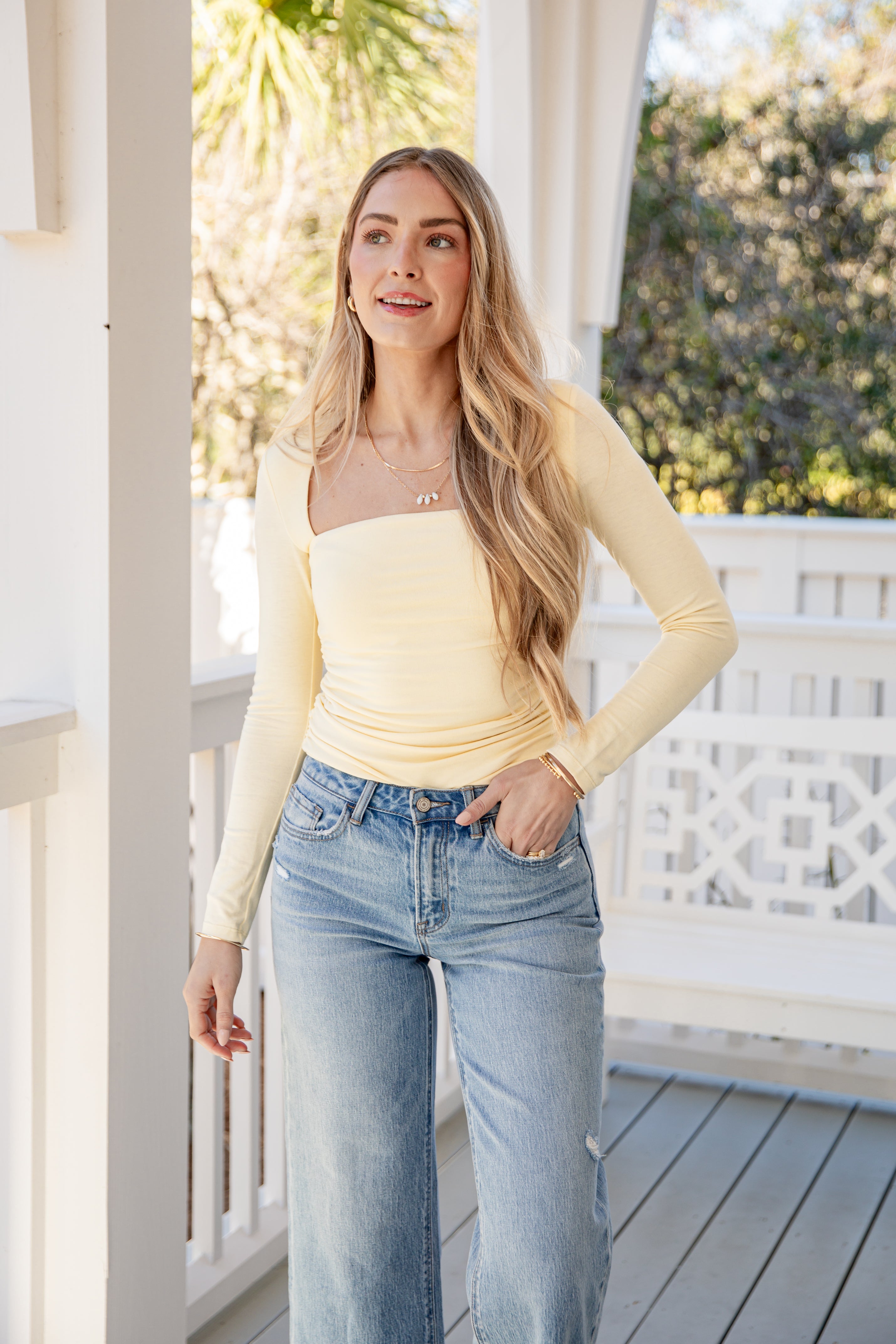 Woman wearing a yellow top and blue jeans standing on a porch.