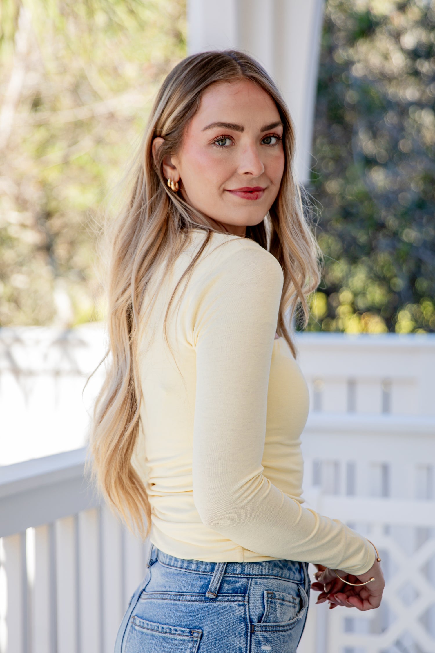 Woman wearing a yellow long-sleeve top and blue jeans on a porch.