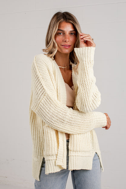 A woman wearing a cream-colored, chunky cable-knit cardigan sweater over a beige top and light-wash blue jeans, standing against a white paneled wall.