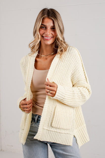 A woman wearing a cream-colored chunky knit cardigan over a beige tank top and light blue jeans, standing against a white paneled wall.