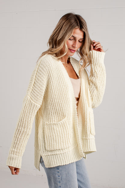 A woman wearing a cream-colored chunky knit cardigan with front pockets, styled over a light beige top and blue jeans, standing against a white paneled wall.