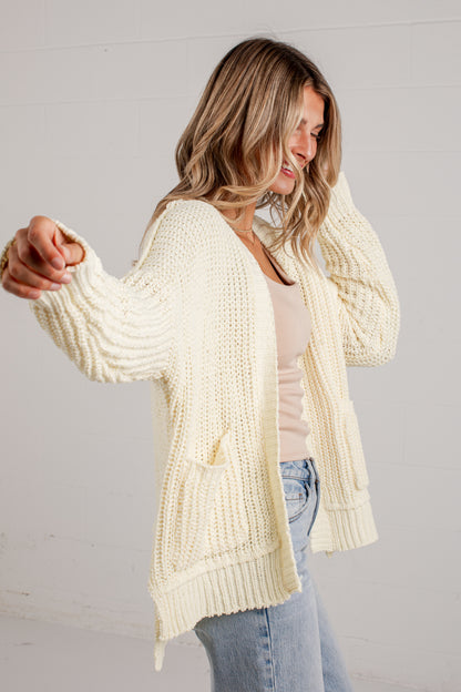A woman wearing a cream cable-knit open-front cardigan over a light pink top and blue jeans, posing against a white paneled wall.