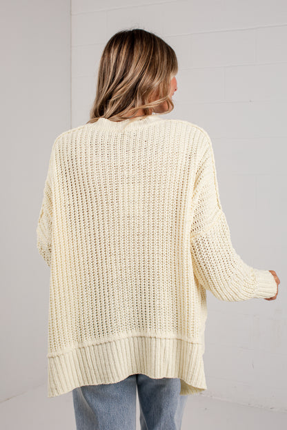 A woman viewed from behind wearing an oversized cream-colored open-knit sweater and light blue jeans, standing against a white paneled wall.