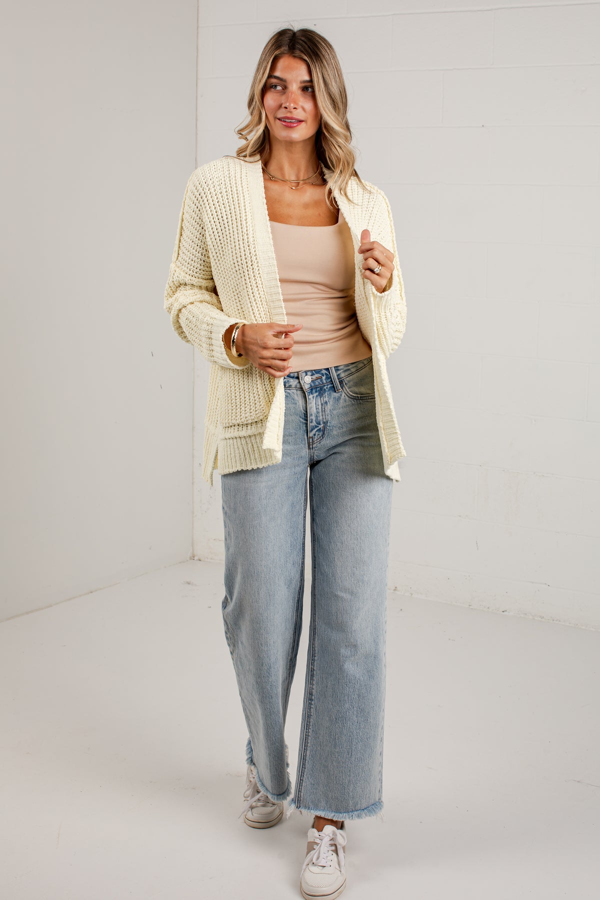 A woman modeling a cream-colored, open-front waffle-knit cardigan over a beige tank top, paired with light-wash wide-leg jeans and white sneakers, standing against a white studio wall.