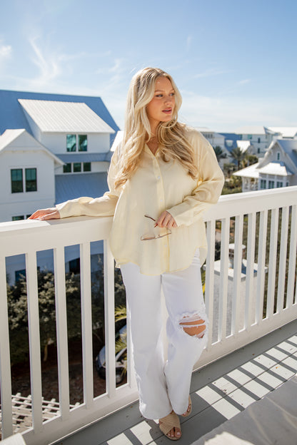 Woman in a light yellow blouse and white pants standing on a balcony with a coastal view.