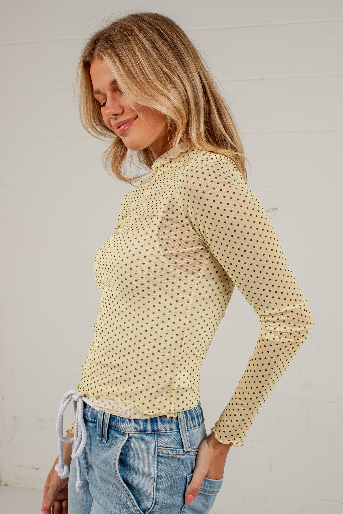 A woman wearing a pale yellow long-sleeve top with a small black polka dot pattern, paired with light blue jeans, standing against a white brick wall.